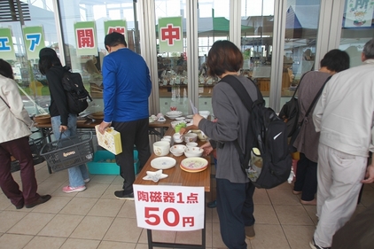 無償の陶磁器ブースを大人複数人が眺めている。1点50円と表記されている。