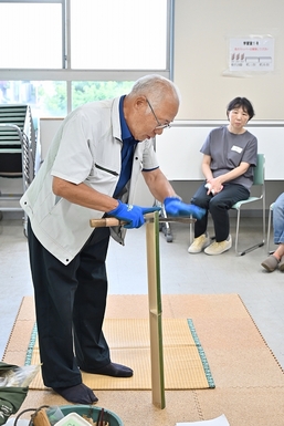 講師の男性が竹をのこぎりできっている。それを参加者がみている。
