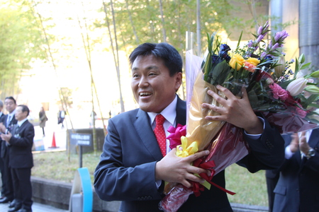 小野塚勝俊市長が笑顔で玄関の前で大きな花束を抱えている